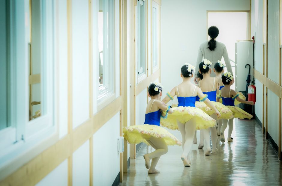 Young ballerinas in tutus follow their instructor through a bright hallway, preparing for dance class.