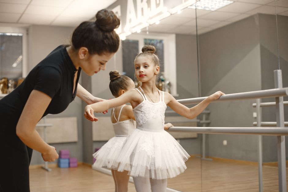 A young girl practices ballet at the barre with guidance from her dance instructor in a studio.