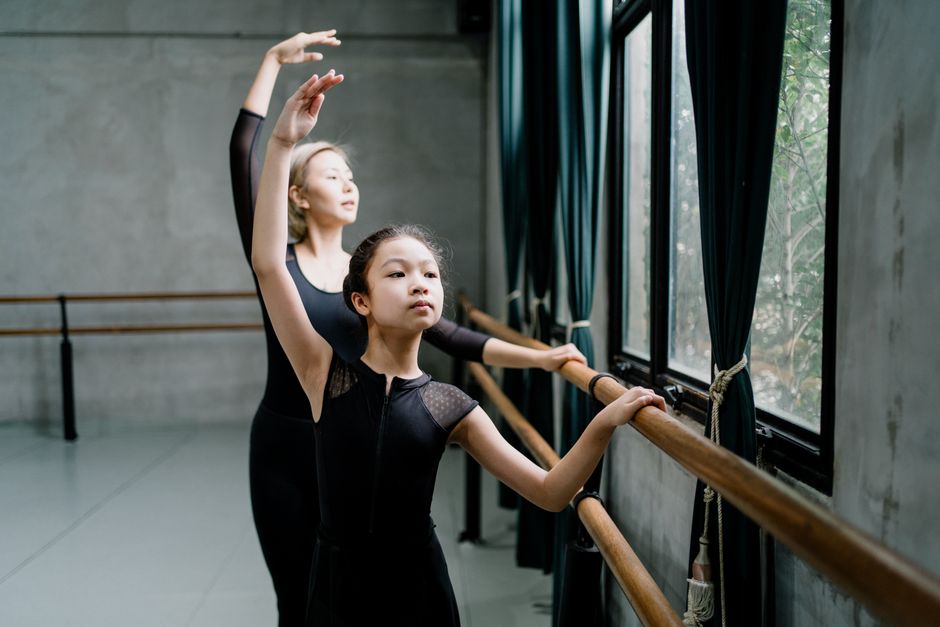 Fit Asian woman and girl ballerinas in black leotards practicing ballet dance while standing near barre with raised arms
