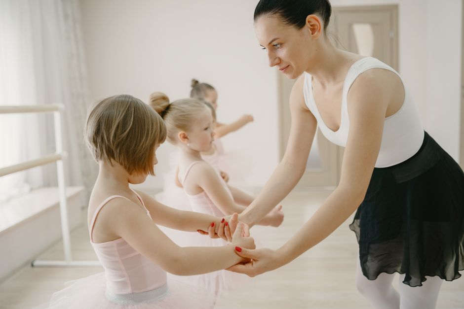 Female ballet teacher guiding young girls in a dance class setting.